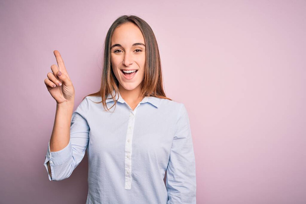 Young beautiful businesswoman wearing elegant shirt standing over isolated pink background showing and pointing up with finger number one while smiling confident and happy. - Foto, imagen