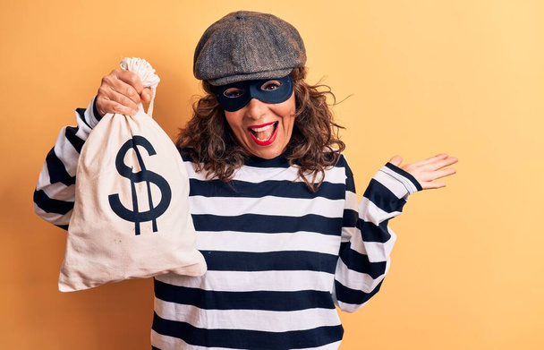 Mujer ladrona morena de mediana edad con máscara y gorra sosteniendo bolsa con símbolo del dólar celebrando el logro con sonrisa feliz y expresión ganadora con la mano levantada
 - Foto, Imagen