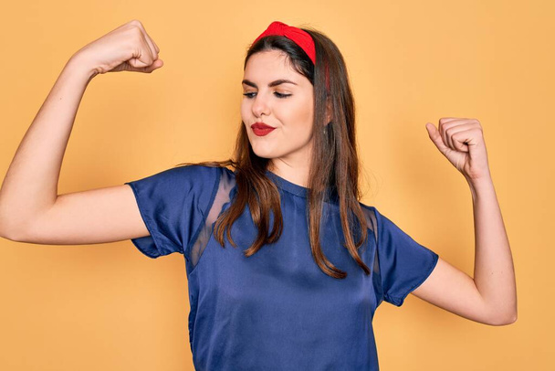 Young beautiful brunette woman wearing red lips over yellow background showing arms muscles smiling proud. Fitness concept. - Fotoğraf, Görsel