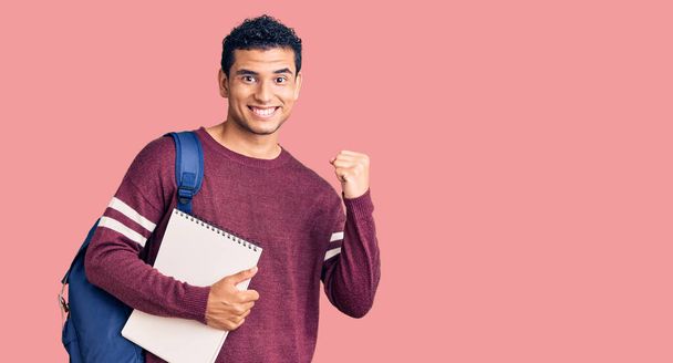 Hispanic handsome young man wearing student backpack and notebook screaming proud, celebrating victory and success very excited with raised arms  - Photo, Image