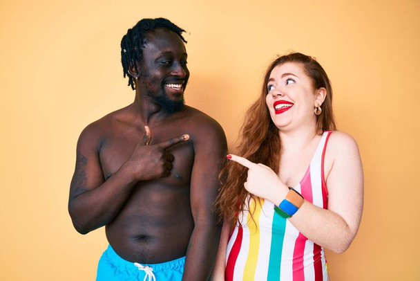 Couple Interracial portant des maillots de bain joyeux avec un sourire du visage pointant avec la main et le doigt sur le côté avec une expression heureuse et naturelle sur le visage  - Photo, image