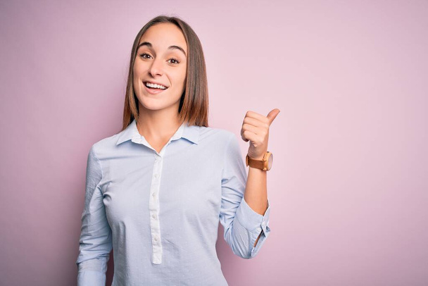 Young beautiful businesswoman wearing elegant shirt standing over isolated pink background smiling with happy face looking and pointing to the side with thumb up. - Photo, Image