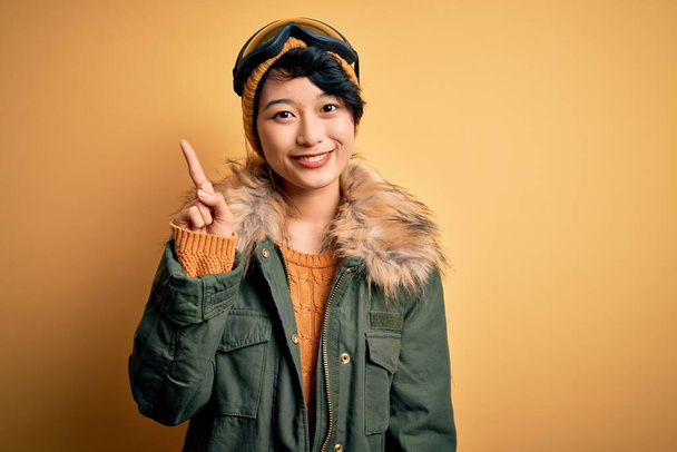 黄色の背景にスキーゴーグルを使用して雪のスポーツウェアを身に着けている美しいアジアのスキー少女は、自信と幸せ笑みを浮かべている間、指のナンバーワンを示すと指摘. - 写真・画像