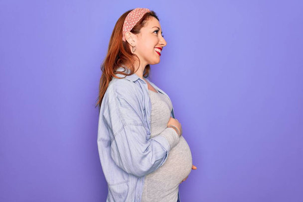 Joven hermosa pelirroja embarazada esperando bebé sobre fondo púrpura aislado mirando a un lado, relajarse pose perfil con cara natural con sonrisa confiada
. - Foto, Imagen