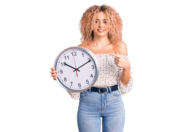 Young blonde woman with curly hair holding big clock smiling happy pointing with hand and finger  - Фото, зображення