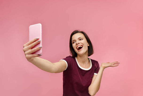 Funny young brunette woman making selfie. Smiling girl wearing t-shirt holding pink smartphone, making faces on camera, posing for selfie isolated on pink background - Фото, изображение