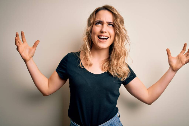 Young beautiful blonde woman wearing casual t-shirt standing over isolated white background crazy and mad shouting and yelling with aggressive expression and arms raised. Frustration concept. - Photo, Image