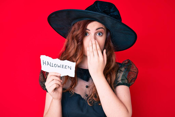 Young beautiful woman wearing witch halloween costume holding paper with halloween message covering mouth with hand, shocked and afraid for mistake. surprised expression  - Photo, Image