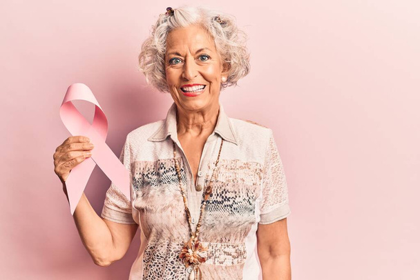 Старша сіро-волохата жінка тримає рожеву ракову стрічку, виглядає позитивно і щасливо стоячи і посміхаючись з впевненою посмішкою, показуючи зуби
  - Фото, зображення