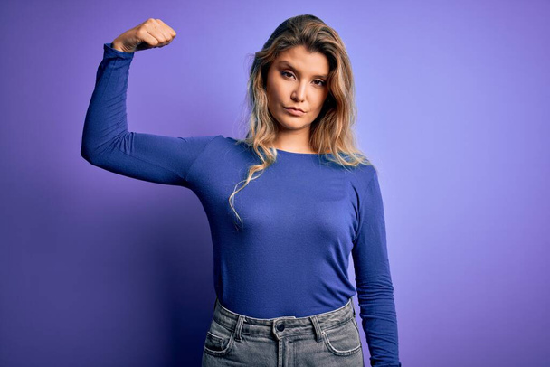 Giovane bella donna bionda indossa casual t-shirt su sfondo viola isolato Persona forte mostrando muscoli del braccio, fiducioso e orgoglioso di potere - Foto, immagini