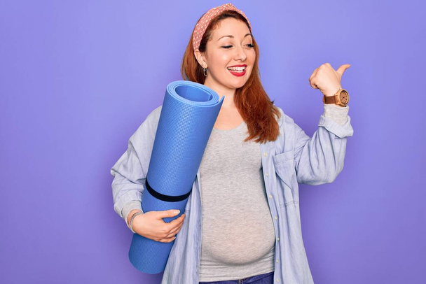 Young beautiful redhead pregnant woman expecting baby holding mat to do prenatal exercise pointing and showing with thumb up to the side with happy face smiling - Φωτογραφία, εικόνα