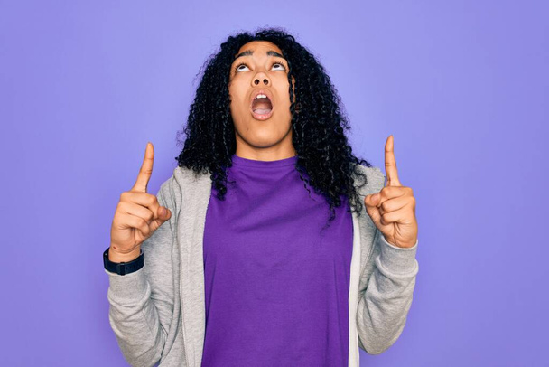 Young african american sporty woman wearing casual sweatshirt over purple background amazed and surprised looking up and pointing with fingers and raised arms. - Photo, Image