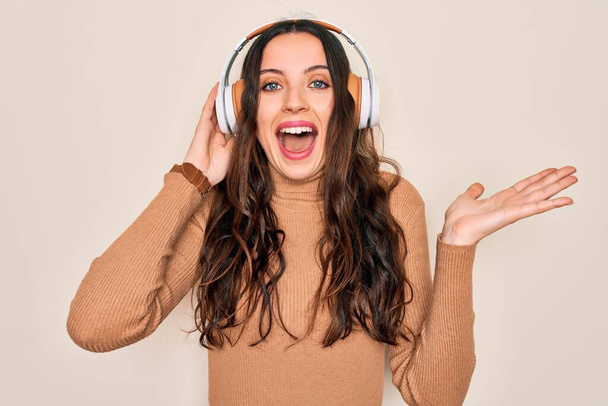 Young beautiful woman with blue eyes listening to music and dancing using headphones very happy and excited, winner expression celebrating victory screaming with big smile and raised hands - Photo, Image