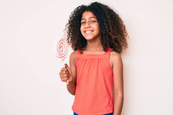 African american child with curly hair holding lollipop looking positive and happy standing and smiling with a confident smile showing teeth  - Foto, immagini