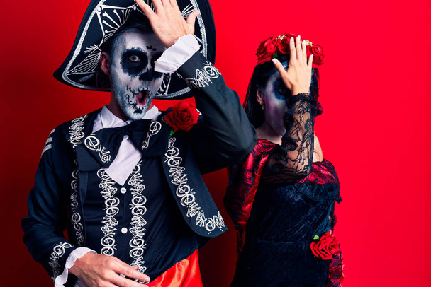 La coppia giovane che indossa il giorno messicano del costume morto sopra rosso sorpreso con mano sulla testa per errore, ricordi l'errore. dimenticato, concetto di cattiva memoria.  - Foto, immagini