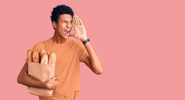 Junger afrikanisch-amerikanischer Mann hält Papiertüte mit Brot in der Hand und schreit laut Seite an Seite mit der Hand auf dem Mund. Kommunikationskonzept.  - Foto, Bild