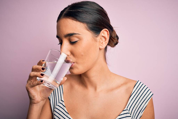 Νεαρή όμορφη μελαχρινή γυναίκα πίνοντας ποτήρι υγιεινό νερό για να δροσιστεί στέκεται πάνω από απομονωμένο ροζ φόντο - Φωτογραφία, εικόνα