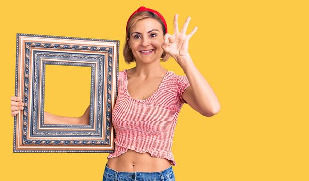 Young blonde woman holding empty frame doing ok sign with fingers, smiling friendly gesturing excellent symbol  - Photo, Image