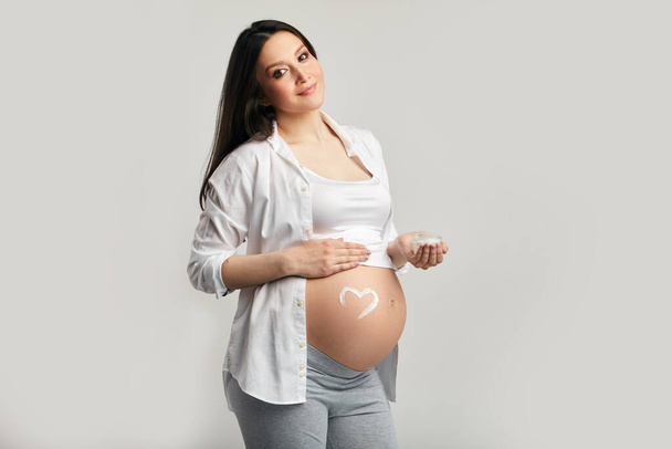 Loving pregnant woman looking at camera while posing in studio against grey wall - Fotografie, Obrázek