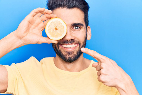 Молодий красивий чоловік з бородою тримає шматочок лимона над оком посміхаючись щасливим вказуючи рукою і пальцем
  - Фото, зображення