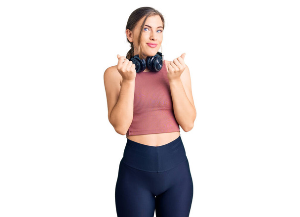 Beautiful caucasian young woman wearing gym clothes and using headphones doing money gesture with hands, asking for salary payment, millionaire business  - Foto, imagen