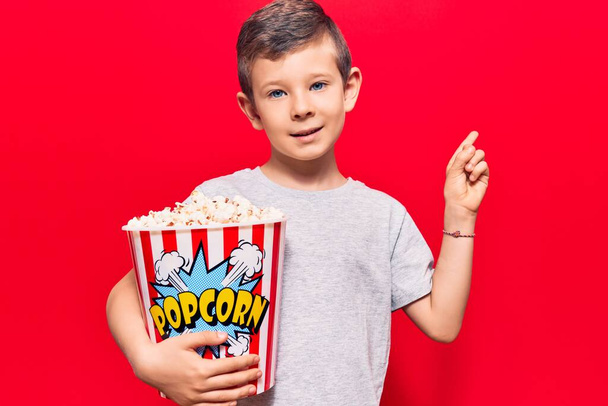 Roztomilé blonďaté dítě držící popcorn s úsměvem šťastný ukazující rukou a prstem na stranu  - Fotografie, Obrázek