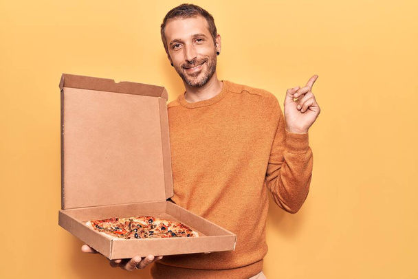 Joven hombre guapo sosteniendo la caja de pizza de entrega sonriendo feliz señalando con la mano y el dedo a un lado  - Foto, imagen
