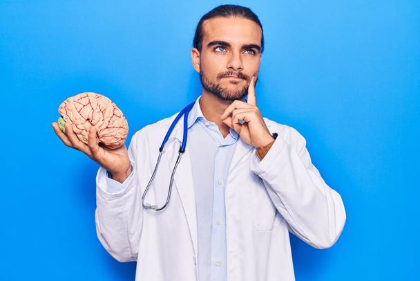 Young handsome man wearing doctor coat holding brain serious face thinking about question with hand on chin, thoughtful about confusing idea  - Foto, Bild