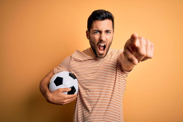 Schöner Mann mit Bart, der Fußball spielt, Ball über gelbem Hintergrund hält und frustriert in die Kamera zeigt, wütend und wütend auf dich - Foto, Bild