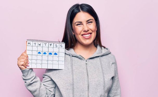 Young beautiful brunette woman holding weather calendar showing rainy week looking positive and happy standing and smiling with a confident smile showing teeth - Zdjęcie, obraz