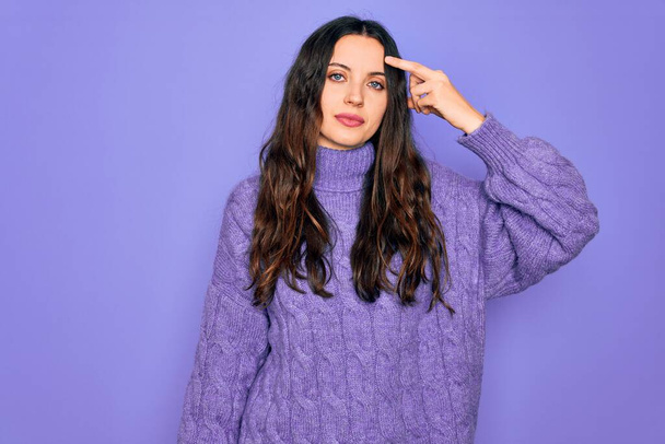 Young beautiful woman wearing casual turtleneck sweater standing over purple background pointing unhappy to pimple on forehead, ugly infection of blackhead. Acne and skin problem - Foto, imagen