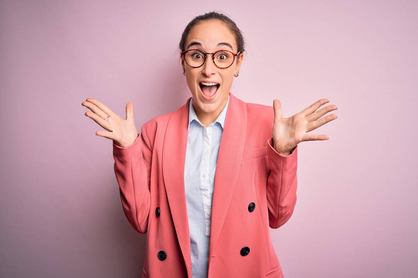 Молодая красивая деловая женщина в пиджаке и очках на изолированном розовом фоне празднует сумасшедший и поражен успехом с поднятыми руками и открытыми глазами, крича взволнованно. Концепция победителя - Фото, изображение