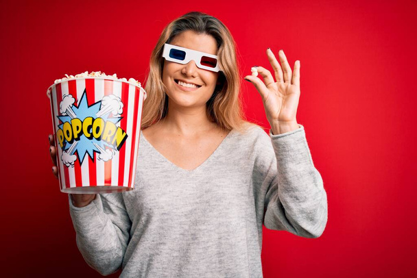 Genç, sarışın bir kadın film izliyor. 3D gözlükler takıyor. Mutlu bir yüzle patlamış mısır yiyor. - Fotoğraf, Görsel