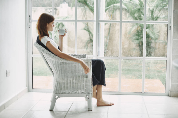 Красивая женщина в кресле у окна с чашкой кофе в руке
  - Фото, изображение