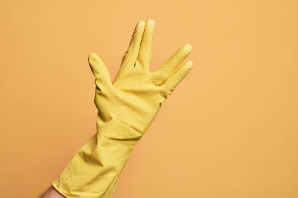 Ręka białego młodzieńca w rękawiczce czyszczącej nad odizolowanym żółtym tłem, salutująca wolkańskiemu salutowi, pokazująca dłoń i palce, dziwaczna kultura - Zdjęcie, obraz