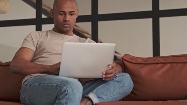 A calm african american man is using his laptop computer sitting on the sofa in living room at home - Metraje, vídeo