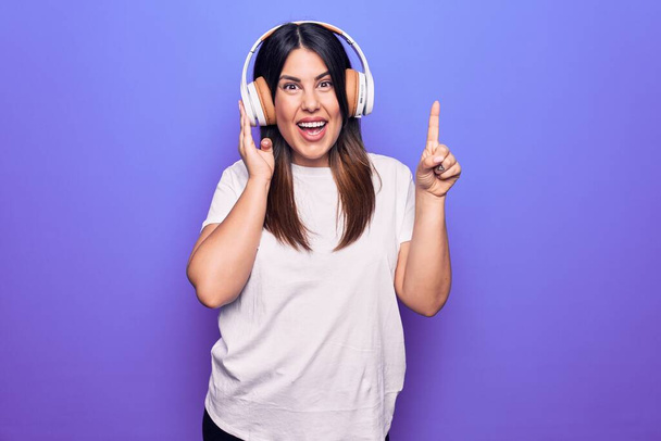 Jeune belle femme brune écoutant de la musique à l'aide d'écouteurs sur fond violet souriant avec une idée ou une question pointant du doigt avec un visage heureux, numéro un - Photo, image