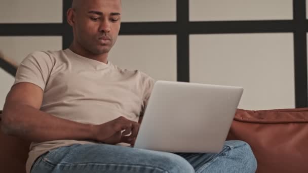A handsome african american man is looking to his laptop sitting on the couch in living room at home - 映像、動画