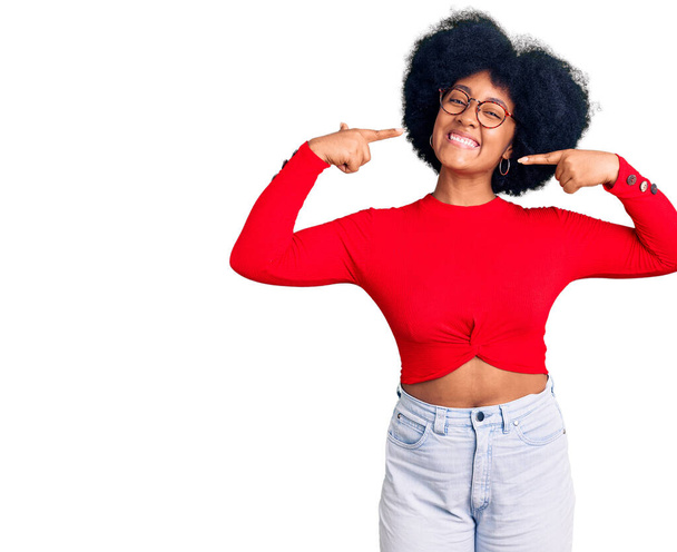 Jeune fille afro-américaine portant des vêtements décontractés et des lunettes souriant joyeuse montrant et pointant du doigt les dents et la bouche. concept de santé dentaire.  - Photo, image