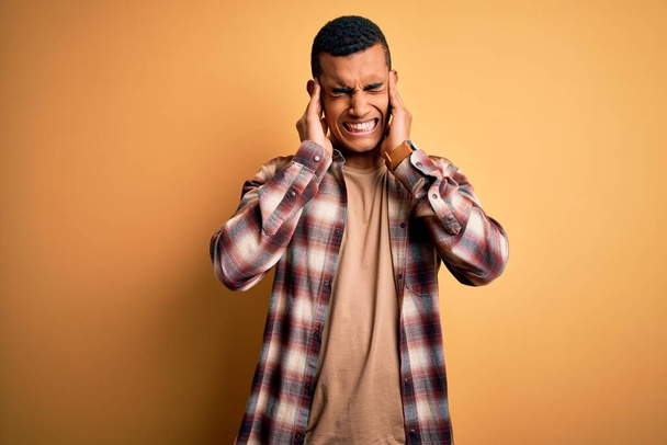 Junger gutaussehender afrikanisch-amerikanischer Mann in lässigem Hemd, das über gelbem Hintergrund steht und die Ohren mit Fingern bedeckt, die genervt sind von dem Lärm lauter Musik. Gehörloses Konzept. - Foto, Bild