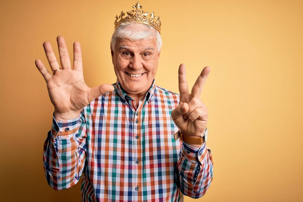 Senior handsome hoary man wearing golden crown of king over isolated yellow background showing and pointing up with fingers number seven while smiling confident and happy. - Fotografie, Obrázek