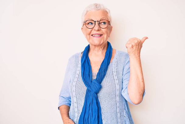 Senior beautiful woman with blue eyes and grey hair wearing casual sweater and scarf pointing thumb up to the side smiling happy with open mouth  - Photo, Image