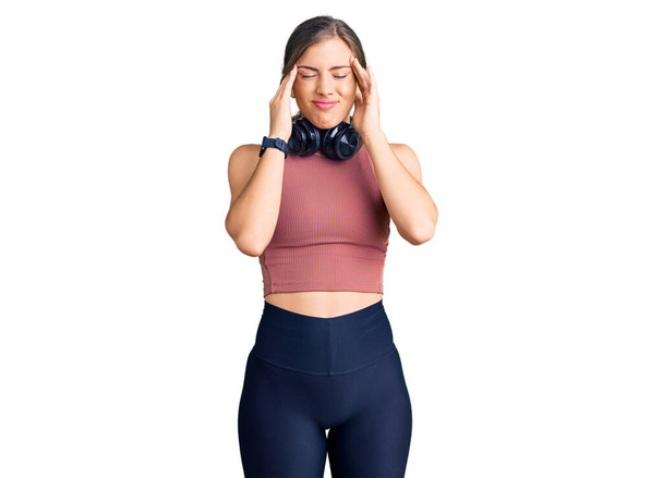 Beautiful caucasian young woman wearing gym clothes and using headphones with hand on head, headache because stress. suffering migraine.  - Фото, изображение