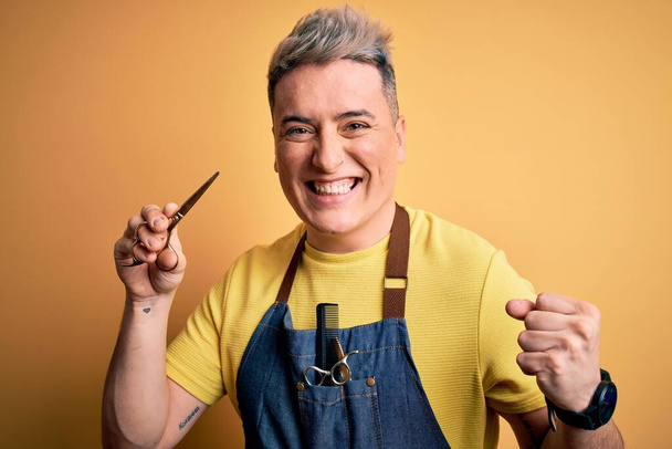 Young modern professional hairdresser man holding scissors over isolated yellow background screaming proud and celebrating victory and success very excited, cheering emotion - Photo, Image