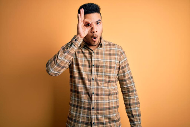 Young handsome man wearing casual shirt standing over isolated yellow background doing ok gesture shocked with surprised face, eye looking through fingers. Unbelieving expression. - Photo, Image