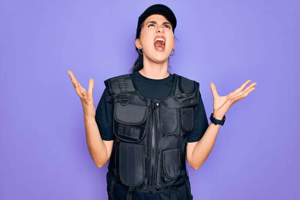 Joven mujer policía vistiendo uniforme chaleco de seguridad a prueba de balas sobre fondo púrpura loco y loco gritando y gritando con expresión agresiva y los brazos levantados. Concepto de frustración
. - Foto, imagen