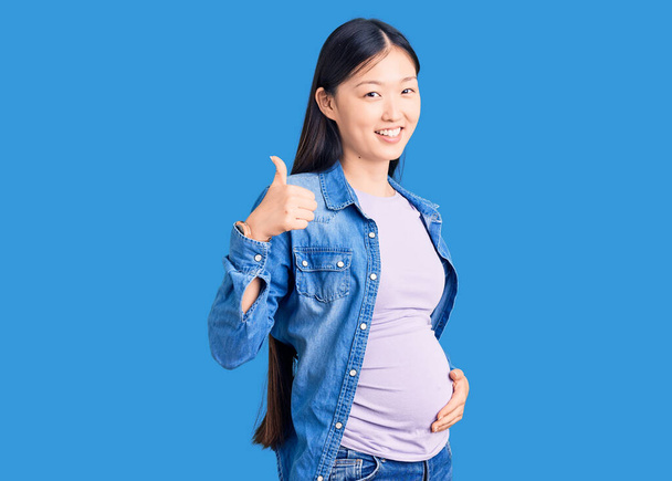 Genç, güzel Çinli hamile bir kadın, bebek beklerken elleriyle mutlu bir jest yapıyor. Kameraya bakıp başarıyı gösteren ifade.  - Fotoğraf, Görsel