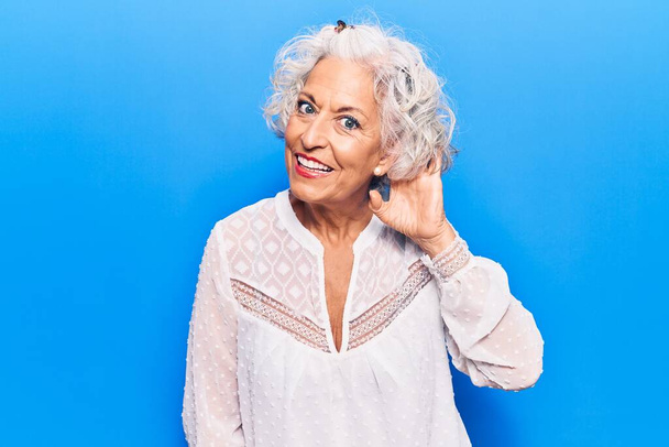 Donna anziana dai capelli grigi che indossa abiti casual sorridendo con mano sull'orecchio ascoltando e ascoltando voci o pettegolezzi. concetto di sordità.  - Foto, immagini