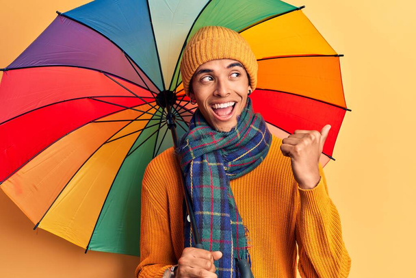 Junger afrikanisch-amerikanischer Mann mit buntem Regenschirm, der den Daumen zur Seite zeigt und glücklich mit offenem Mund lächelt  - Foto, Bild