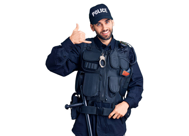 Sakallı, polis üniforması giyen, gülümseyen, el ve parmaklarıyla telefon hareketi yapan, telefonda konuşan genç, yakışıklı bir adam. iletişim kavramları.  - Fotoğraf, Görsel